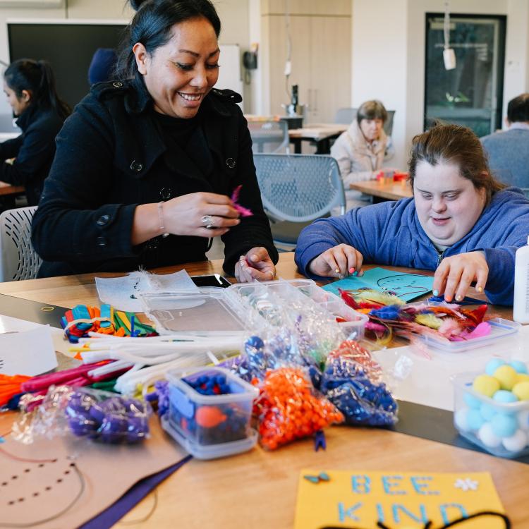 Two people working on a craft project together. There are lots of craft materials on the table and one of the participants is reaching for something